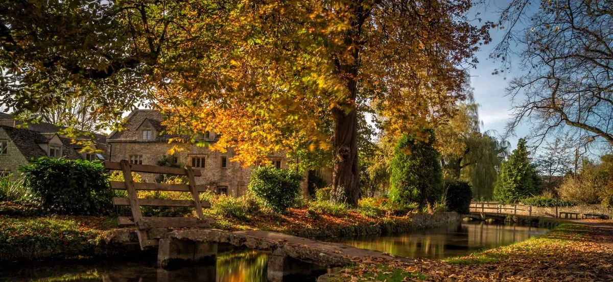 Cotswolds stone cottage and stream framed by trees in autumn color