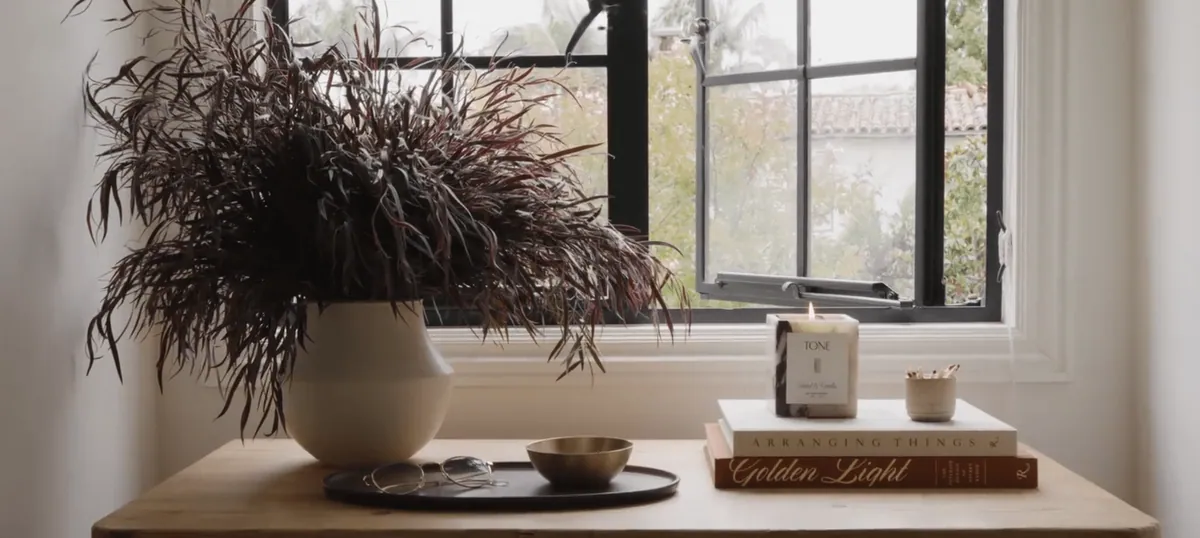Dried plume arrangement in a stone vase on a windowsill beside candles and books