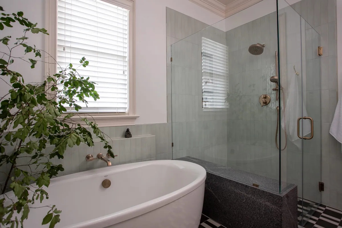 Freestanding soaking tub under a window beside a glass shower with celadon tile and a stone bench
