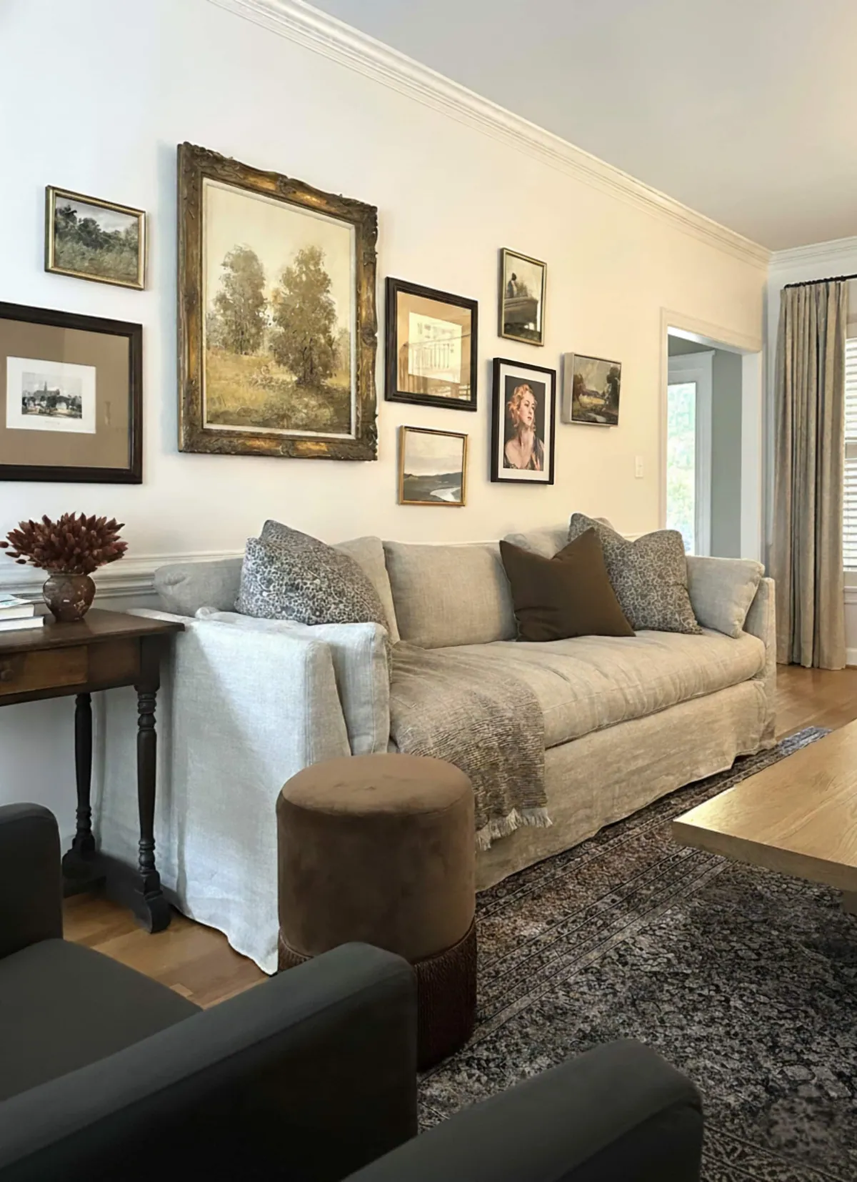 Living room with a linen slipcovered sofa, a gallery wall of framed landscapes in gilt frames and a velvet ottoman