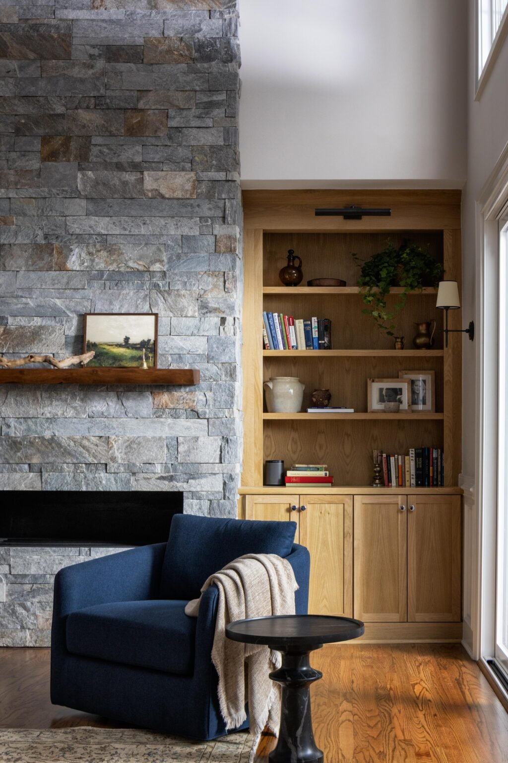 Living room with stone fireplace, wood mantel, built-in oak shelving, and navy armchair
