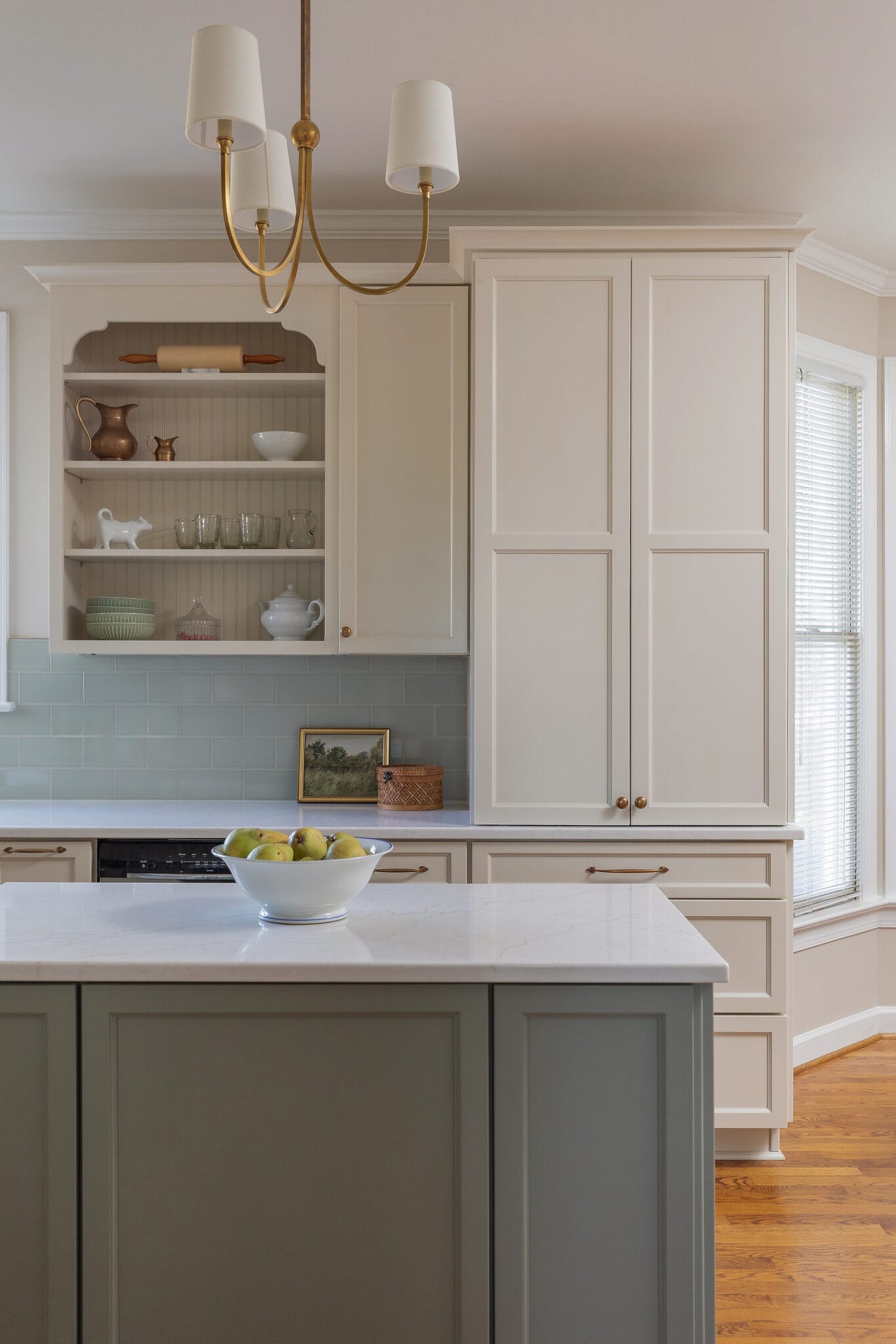 Kitchen with sage green island, cream cabinetry, pale blue glass tile backsplash and brass pendant light