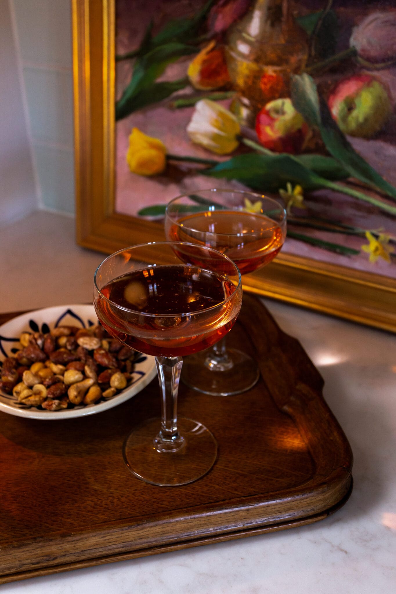 Two cocktails and a bowl of nuts on a wooden tray beneath a floral still life